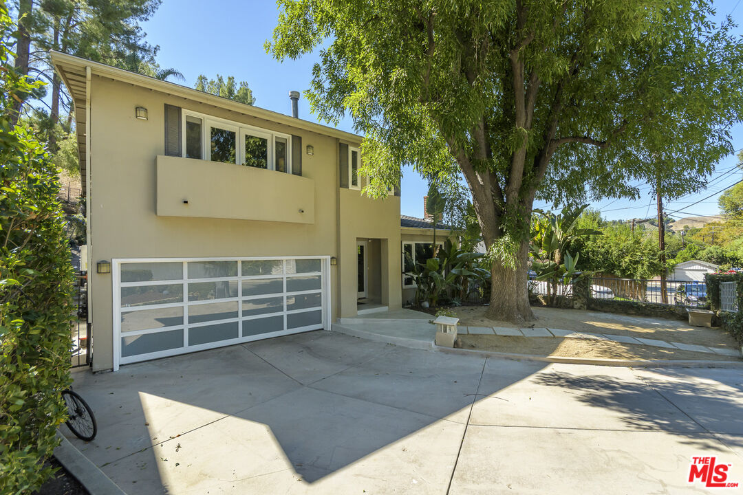 Two-story beige home with a paneled garage door and broad driveway shaded by a mature tree.