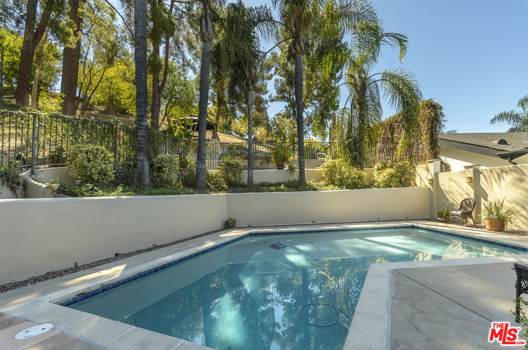 Backyard swimming pool bordered by stone decking, a wall, and tall palm trees.
