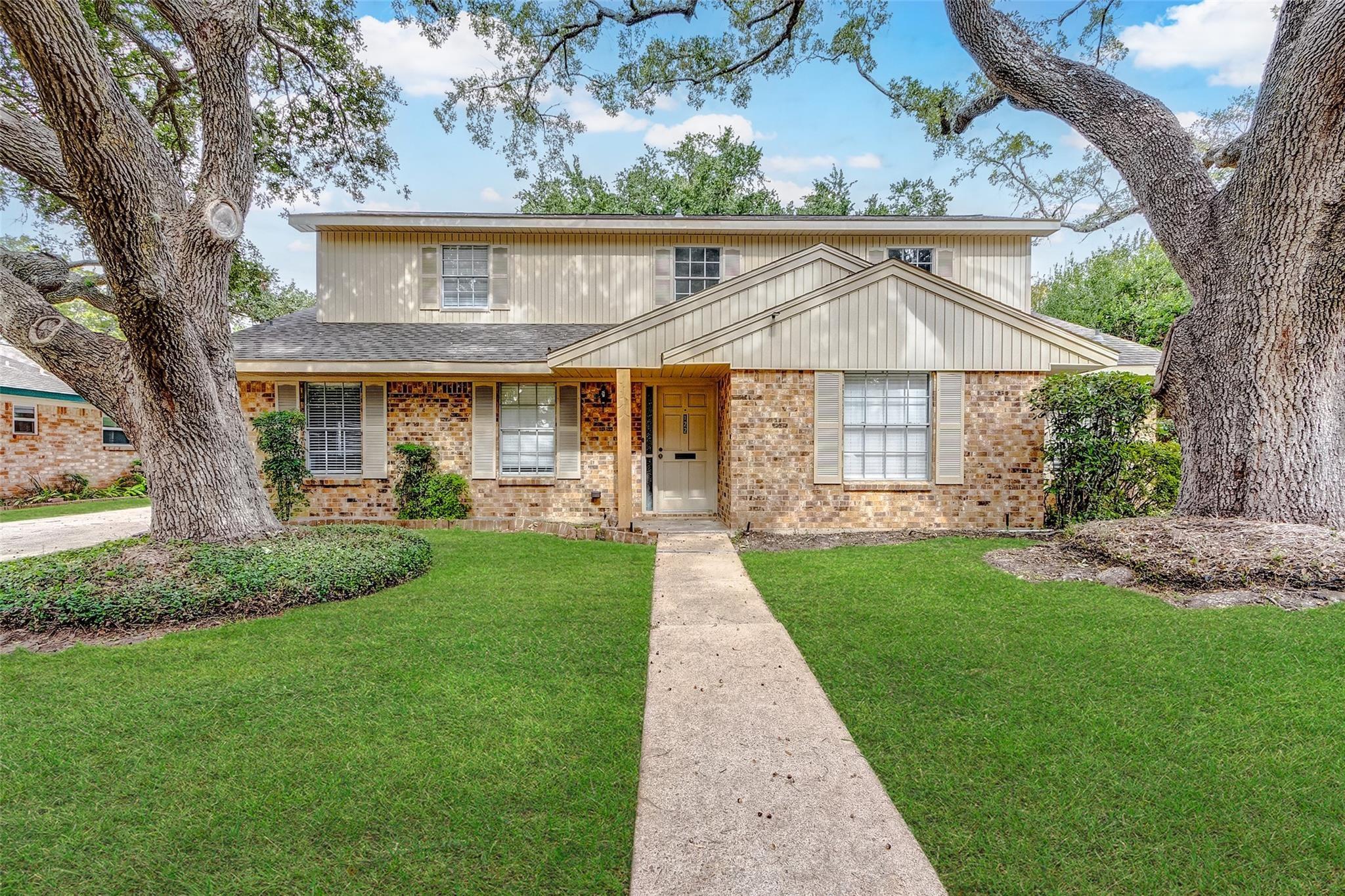 Two-story brick and siding house framed by large oak trees, with a front walkway across the lawn.