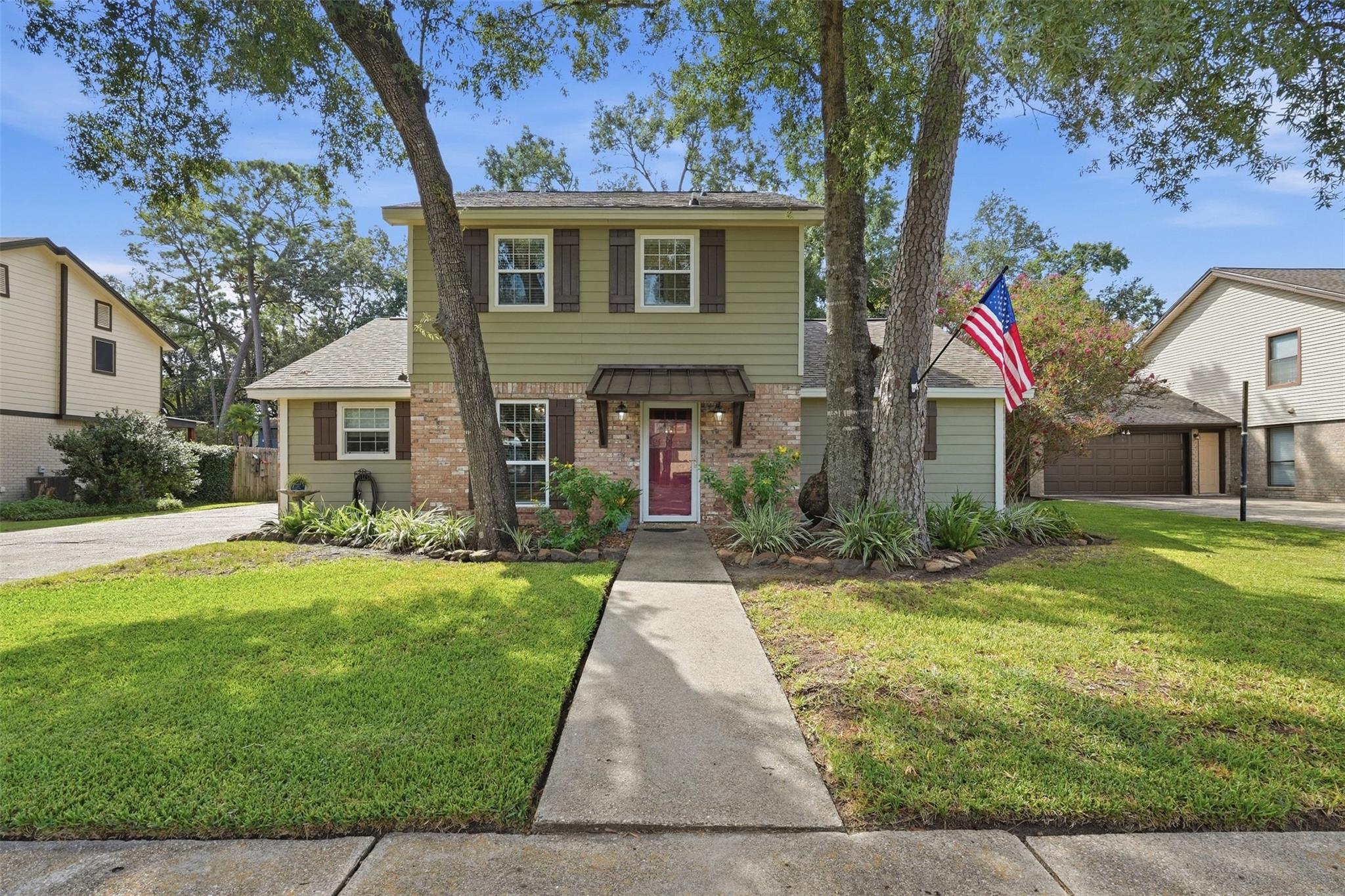 Two-story green-and-brick house with a red front door, American flag, and mature trees.