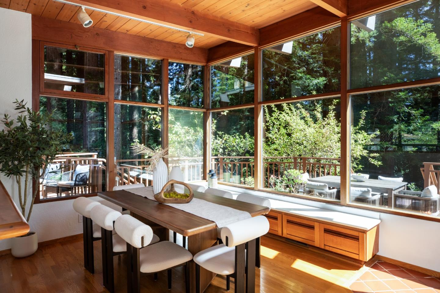 Dining room with a long wooden table beside floor-to-ceiling windows overlooking a leafy garden.