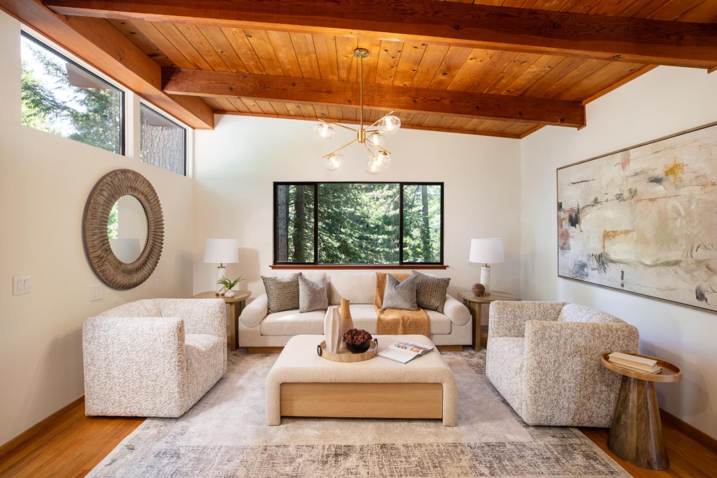 Staged living room with exposed wood ceiling beams, cream seating, and wide windows overlooking trees.