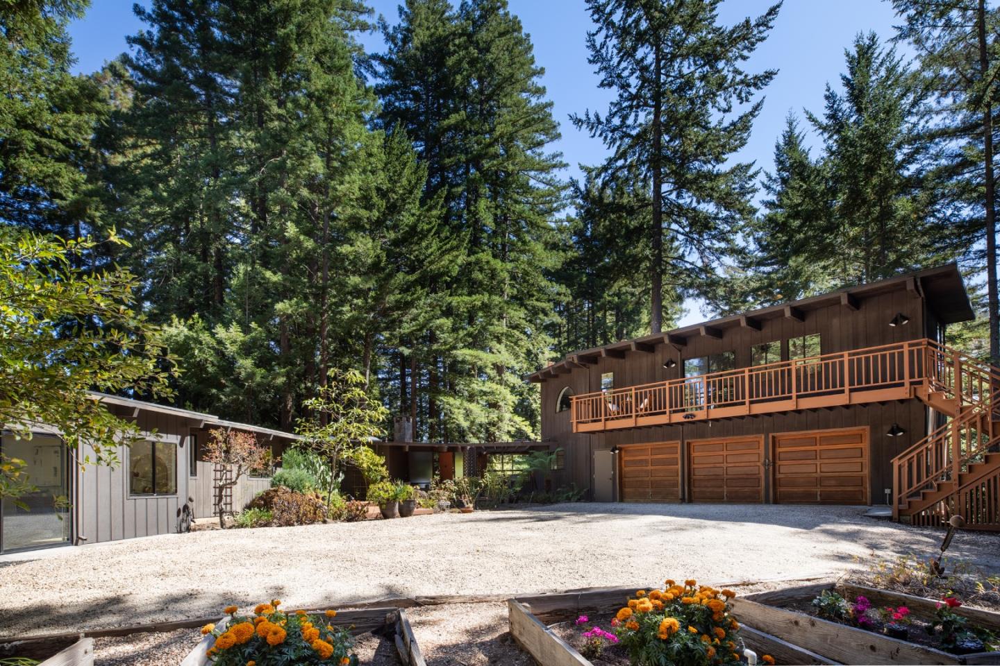 Wood-sided home with a long balcony and three garage doors, surrounded by evergreens and flower beds.
