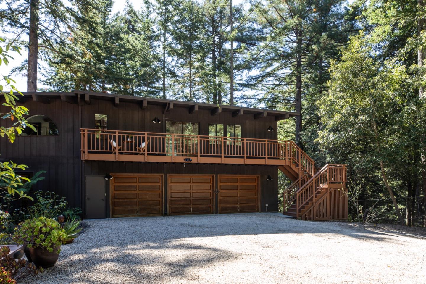 Dark wood house with a raised deck, exterior staircase, and three garage doors among tall trees.