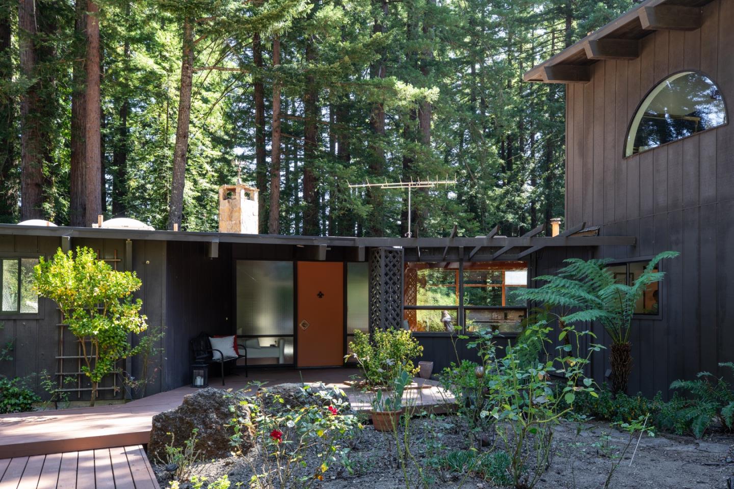 Modern dark-wood home entrance with an orange door, garden deck, and surrounding forest.