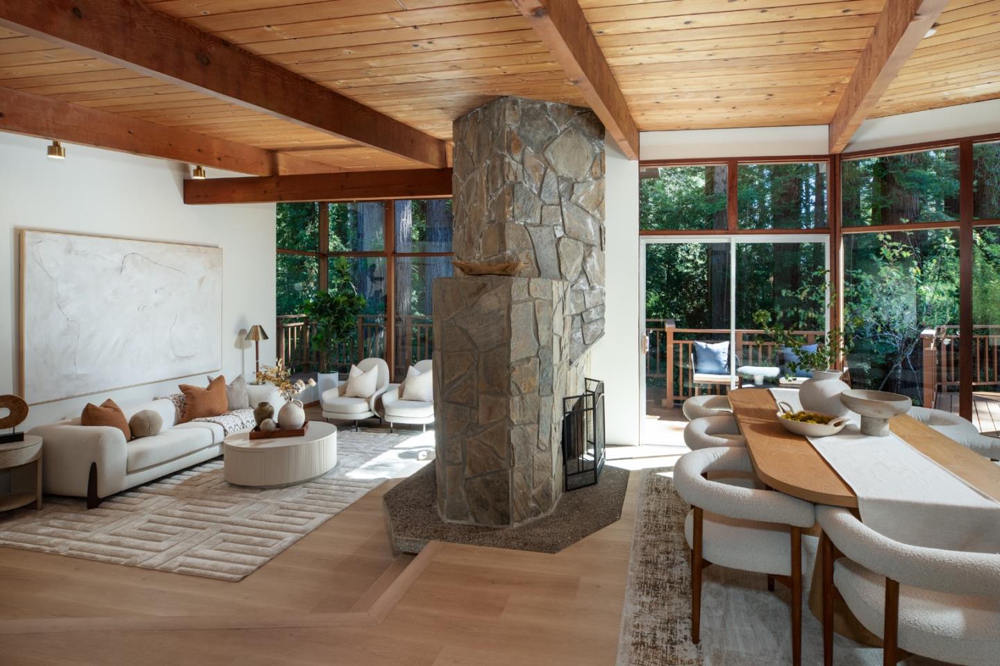 Open living and dining room with a stone fireplace, wood-beam ceiling, and windows facing trees.