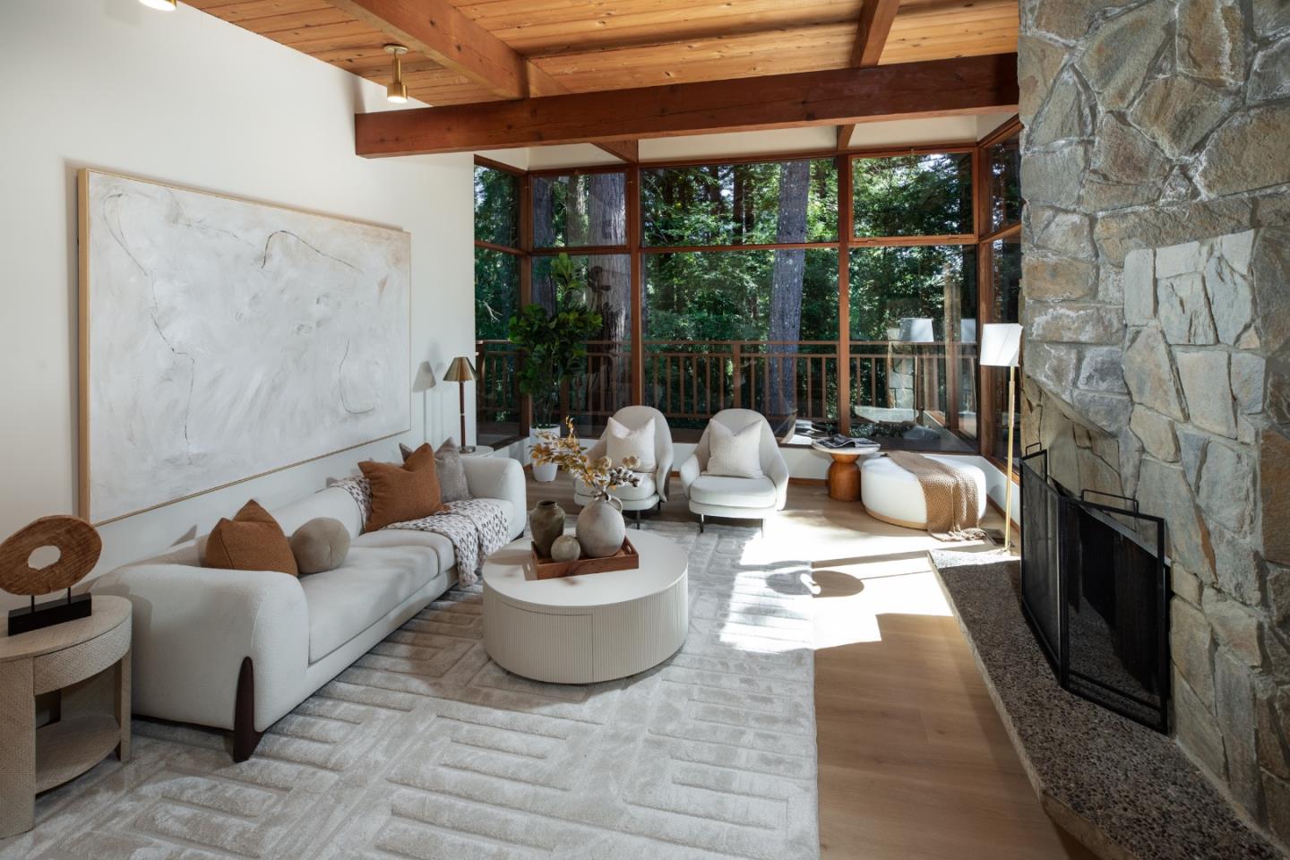 Living room with a stone fireplace, wood ceiling, and large windows overlooking trees.