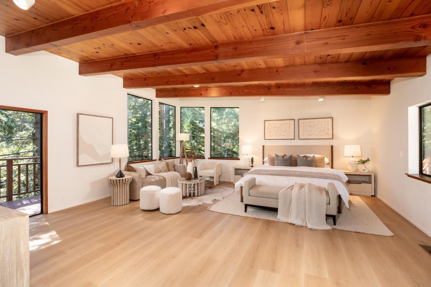 Staged bedroom with exposed timber ceiling beams, a sitting area, and windows overlooking trees.