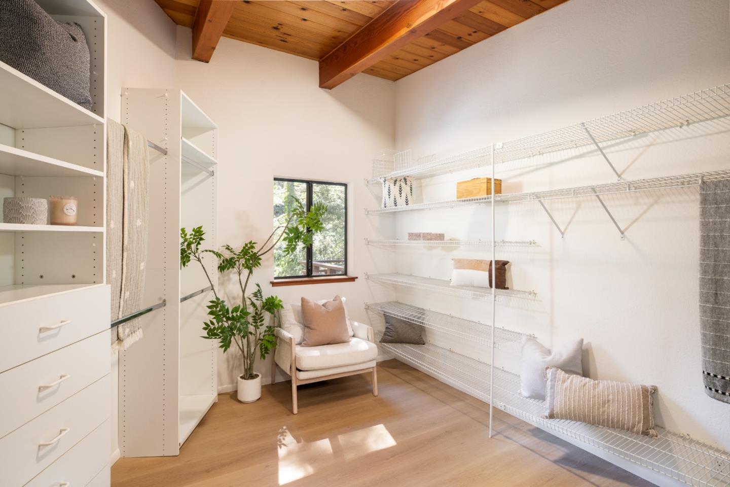 Walk-in closet with white built-ins, open wire shelving, a cushioned chair, and exposed wood ceiling beams.