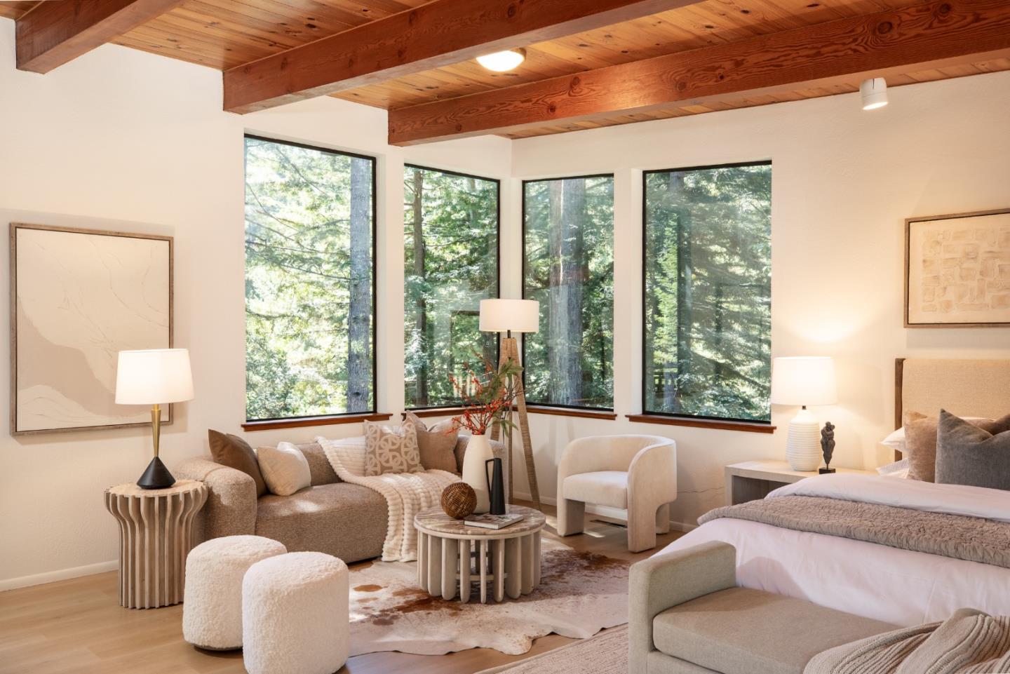 Bedroom with exposed wood beams, a sitting area, and large windows overlooking trees.