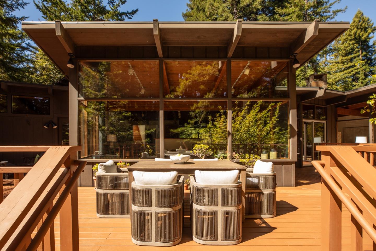 Furnished timber deck with woven dining chairs, a long table, and expansive windows on a modern home.