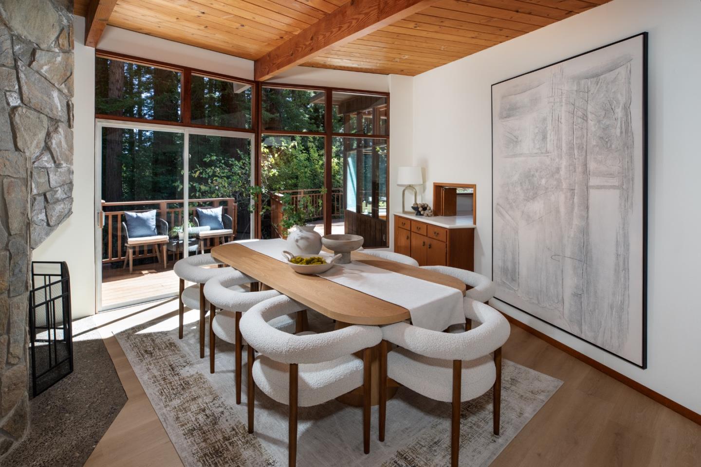 Dining room with a wood-plank ceiling, stone fireplace, and glass doors overlooking a tree-lined deck.