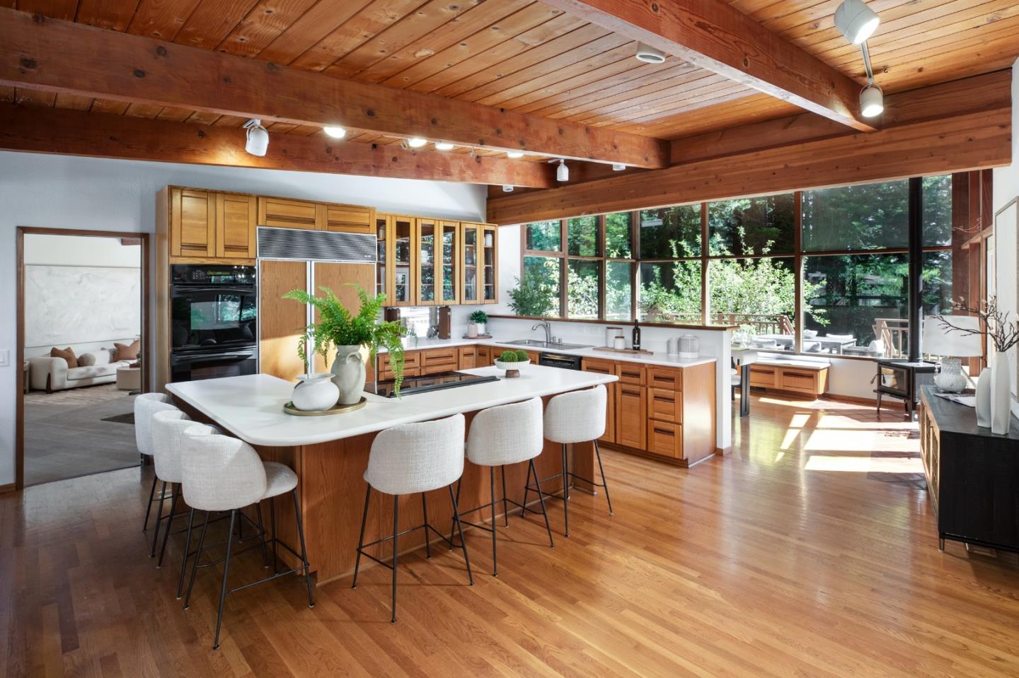 Open-plan kitchen with a large white island, warm wood cabinets, exposed beams, and windows facing greenery.