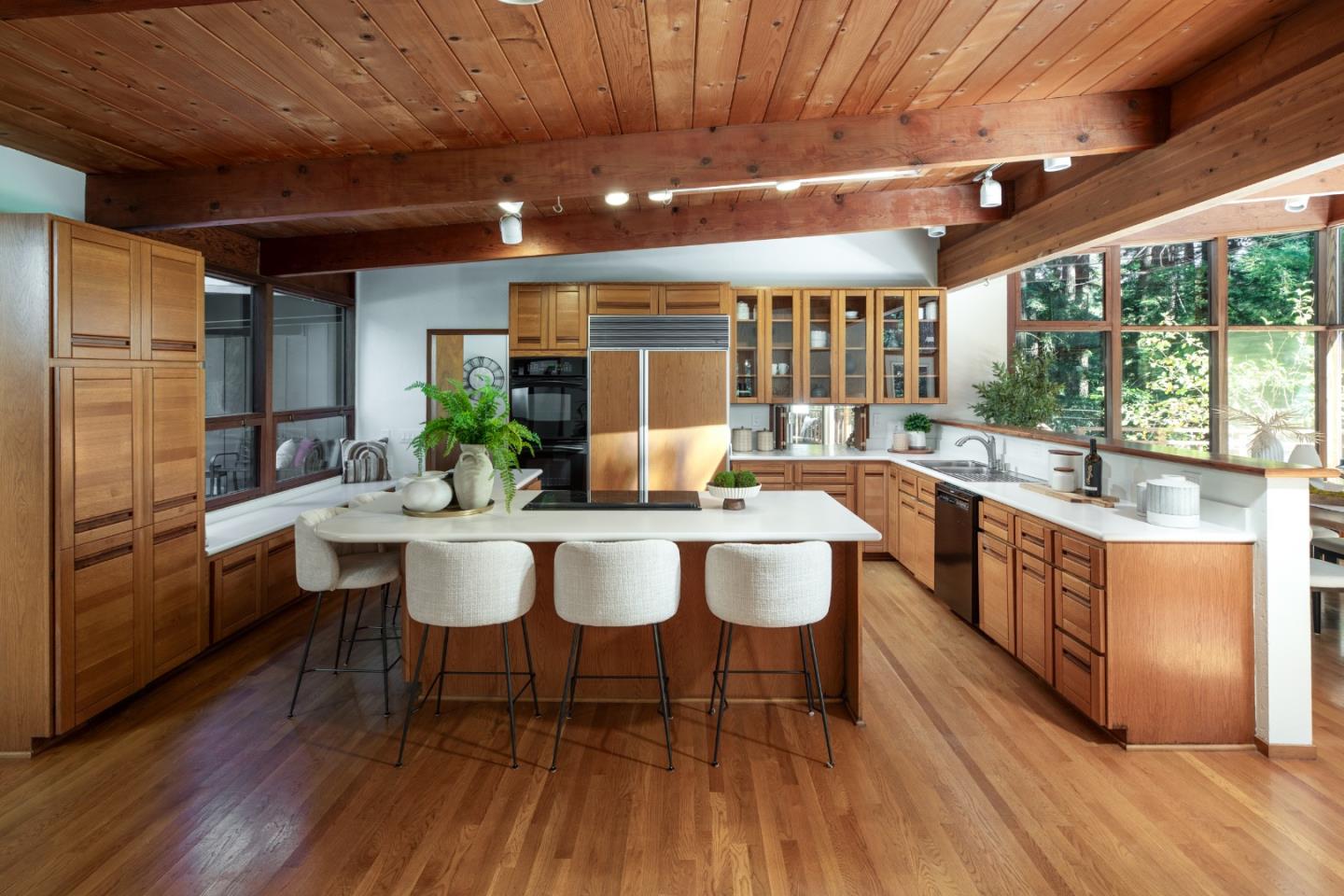 Kitchen with a white island, warm wood cabinetry, exposed beams, and large windows overlooking trees.