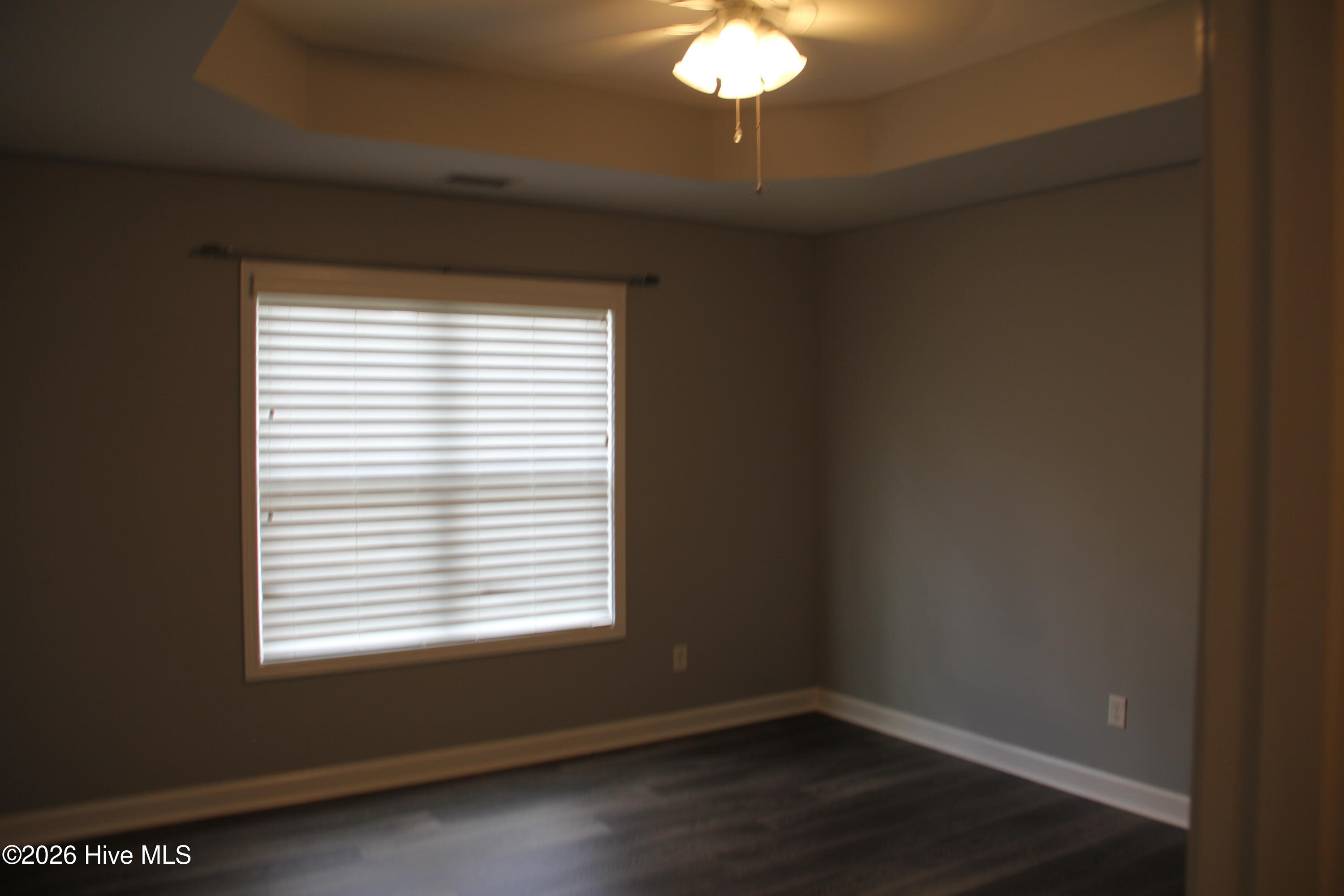 Empty room with dark plank flooring, a window with closed blinds, and a ceiling fan with light.