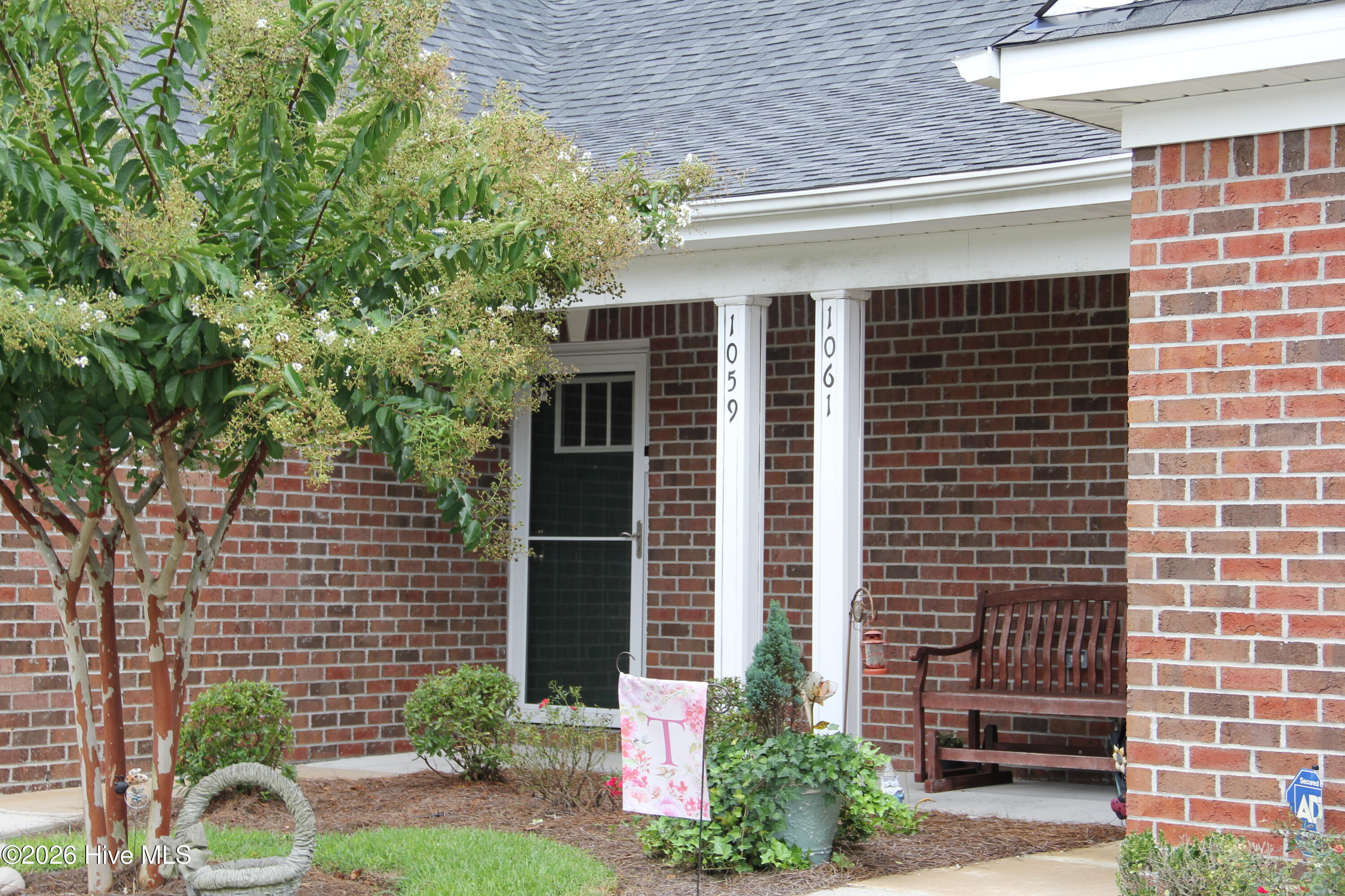 Brick home’s covered porch with white columns, a wooden bench, and flowering greenery.