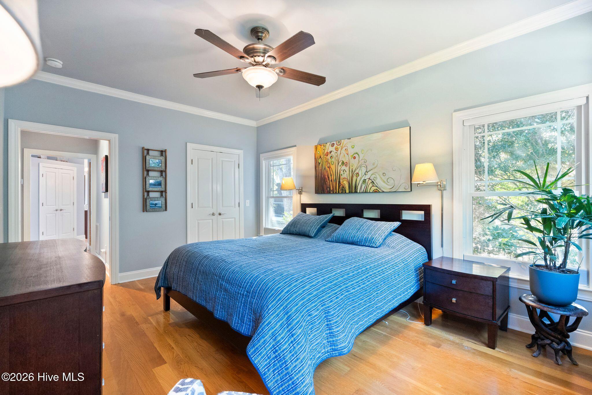Furnished bedroom with a blue-striped bedspread, ceiling fan, hardwood floors, and large windows.