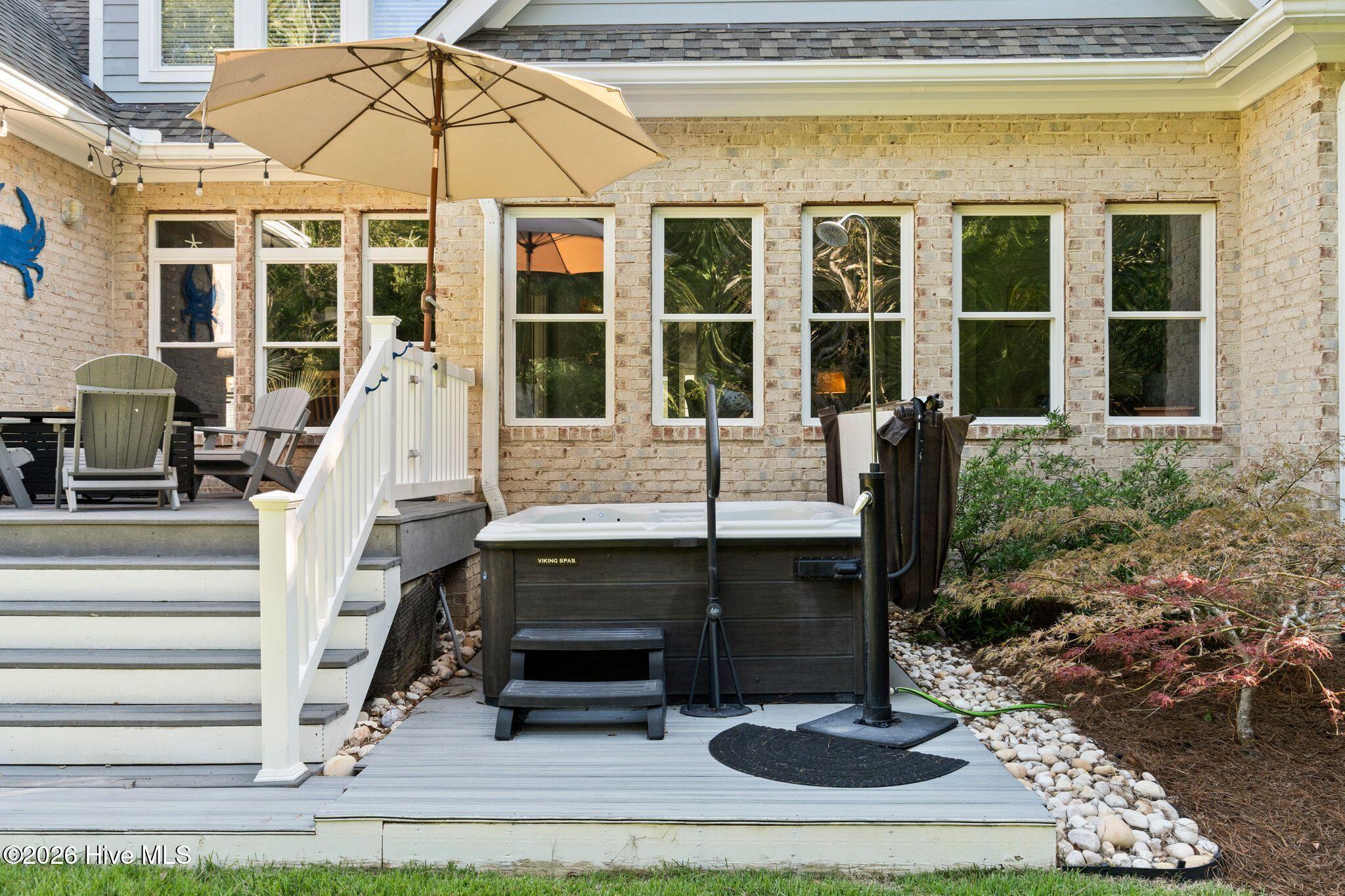 Backyard hot tub beside a brick house, with deck stairs, an umbrella-shaded seating area, and outdoor shower.