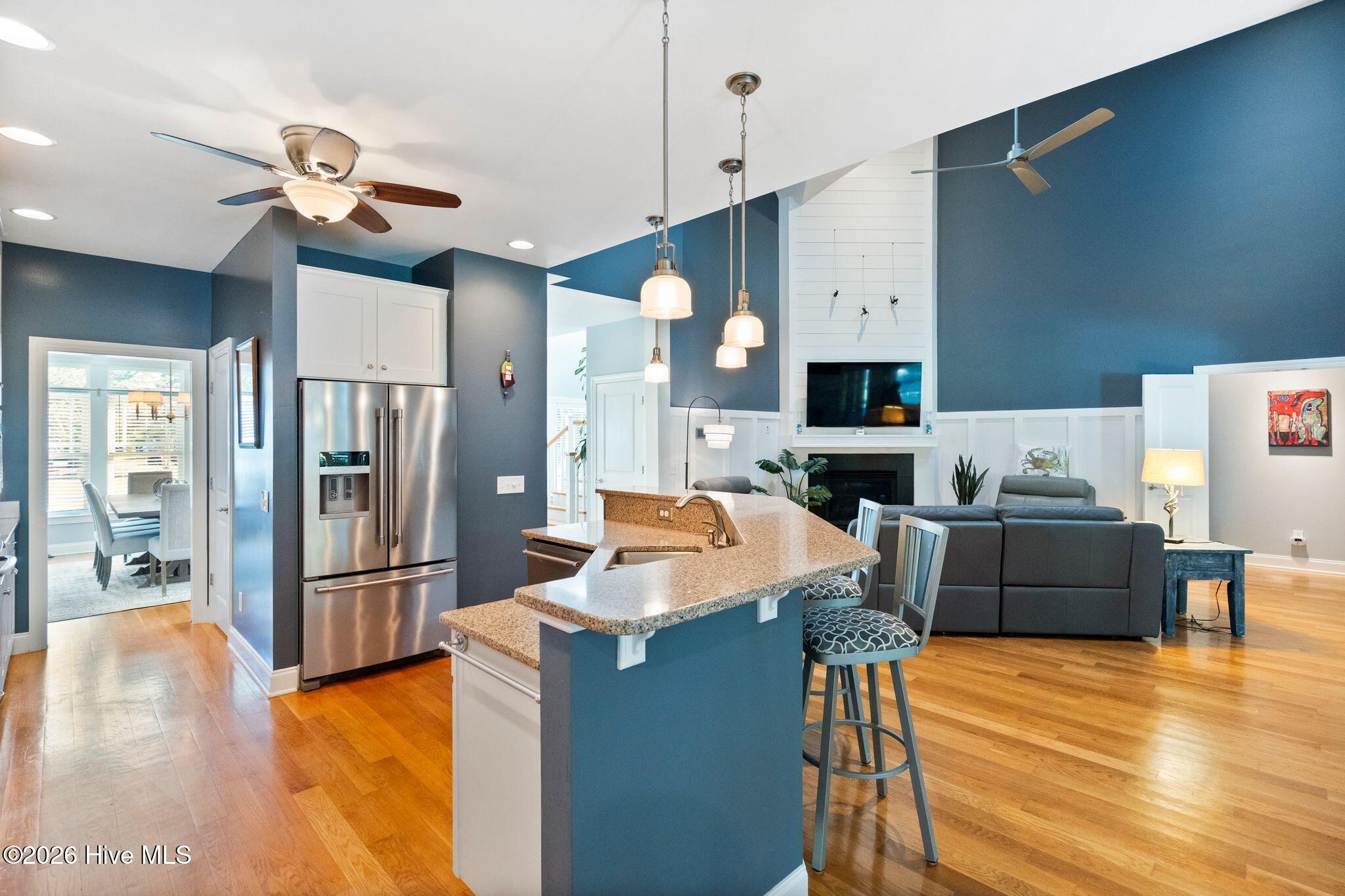 Open kitchen and living room with a granite island, stainless steel refrigerator, hardwood floors, and fireplace.