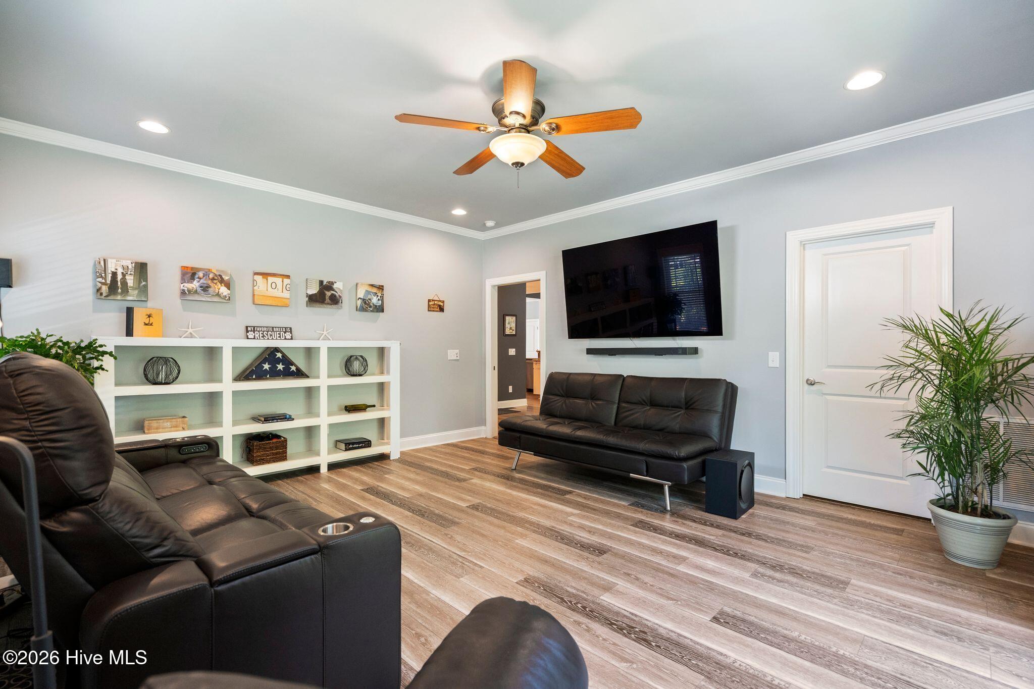 Living room with black leather seating, a wall-mounted TV, white display shelves, and a wood-blade ceiling fan.