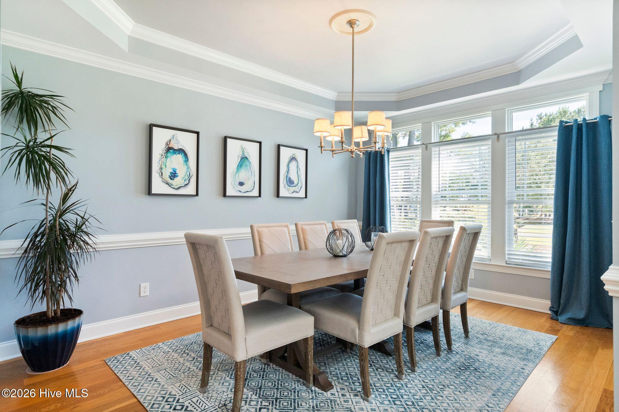 Dining room with a wood table, upholstered chairs, chandelier, and large curtained windows.