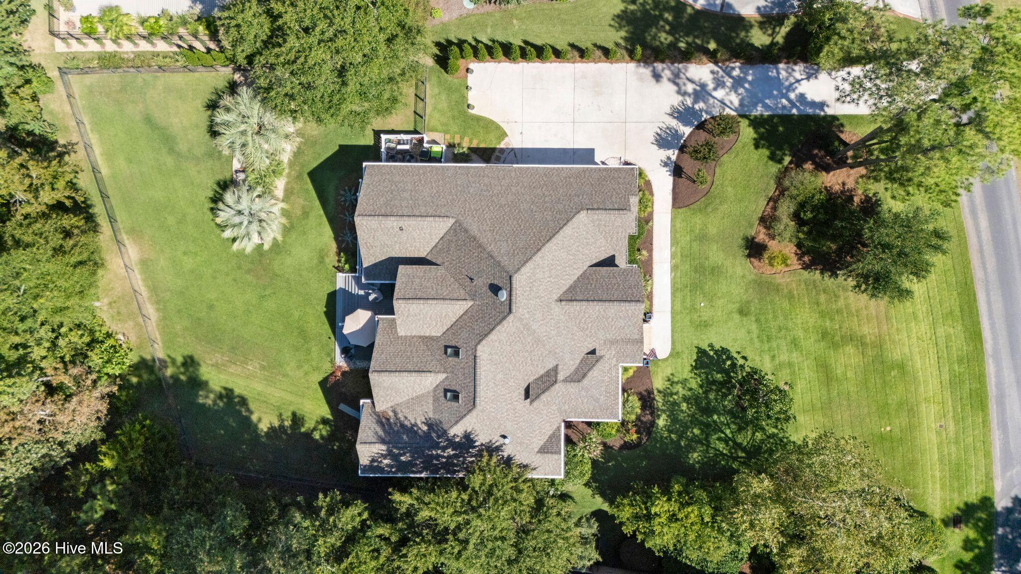 Aerial view of a home with a broad lawn, mature trees, fenced yard, and wide driveway.