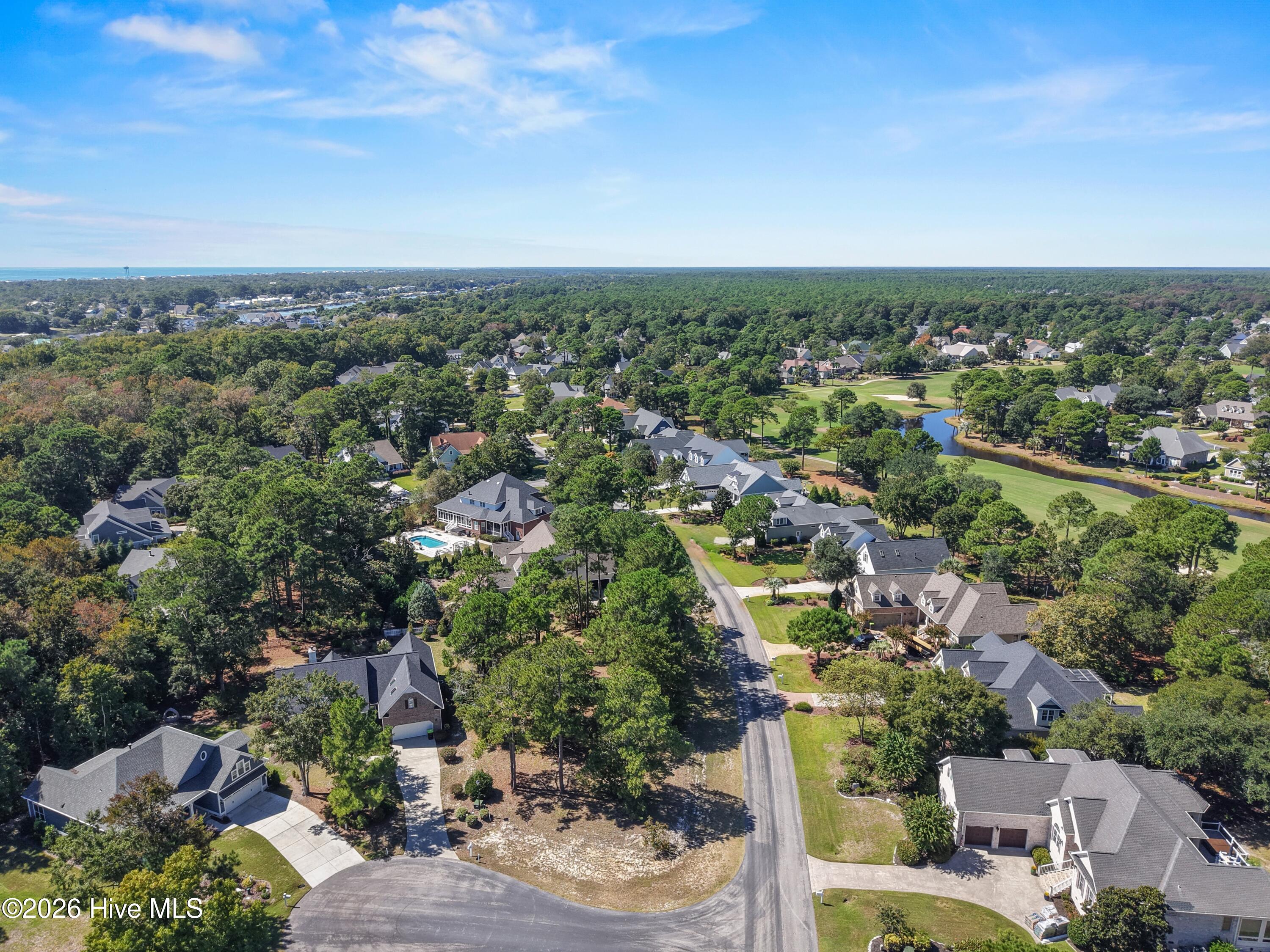 Aerial view of a coastal neighborhood with tree-lined streets, golf course, pond, and distant ocean.