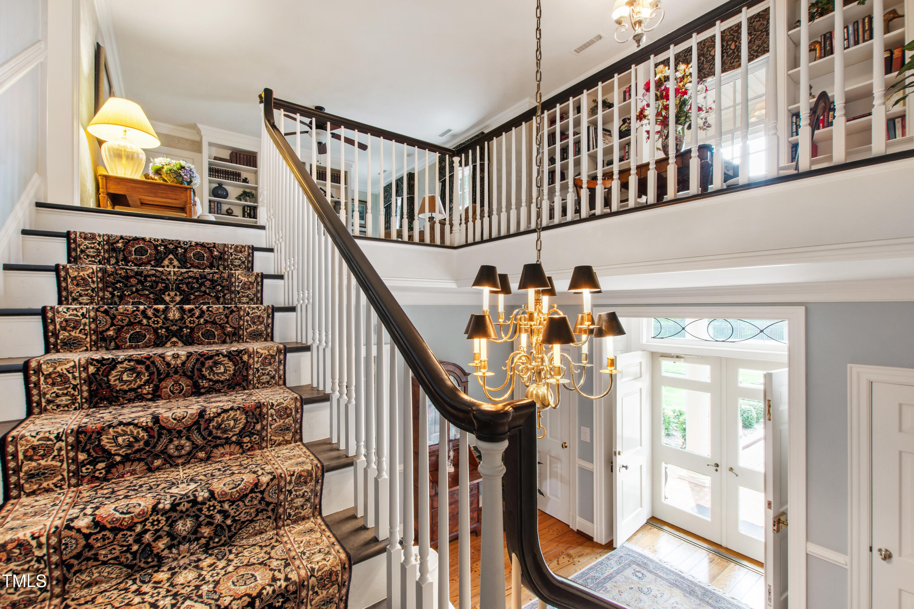 Two-story foyer with a patterned staircase, built-in bookshelves, chandelier, and bright glass-paneled front doors.