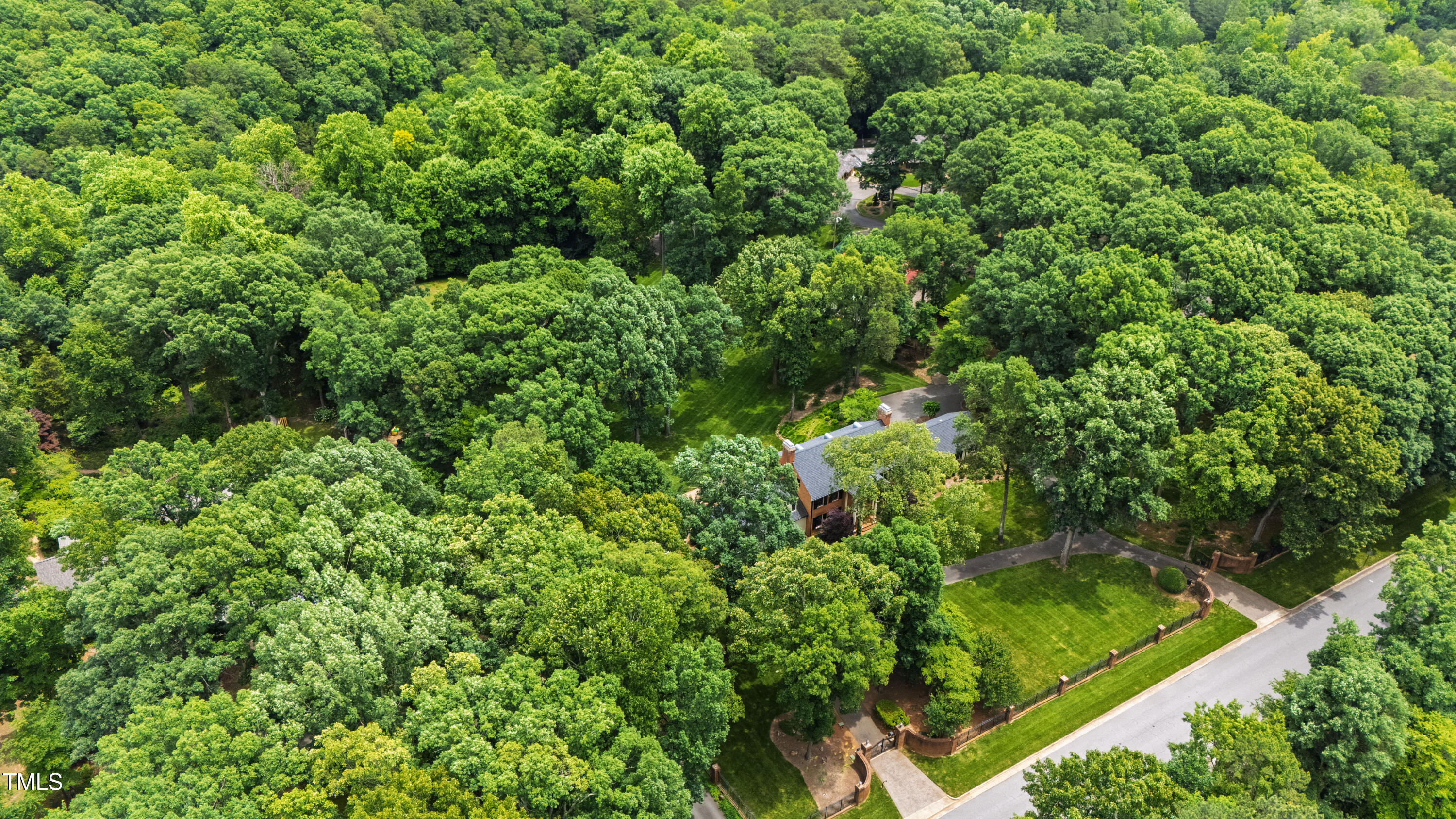 Aerial view of a tree-surrounded home with a broad lawn, curved paths, and a fenced garden.