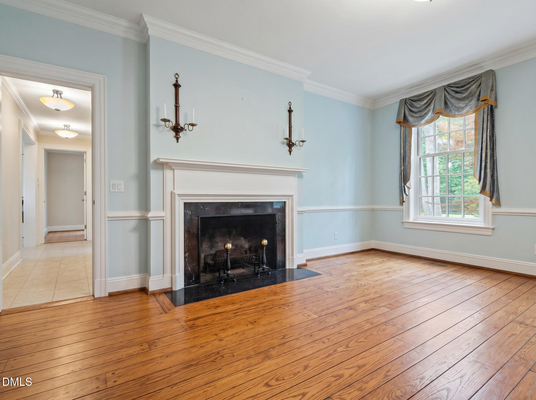Unfurnished living room with a white-trimmed fireplace, pale blue walls, hardwood floors, and a curtained window.