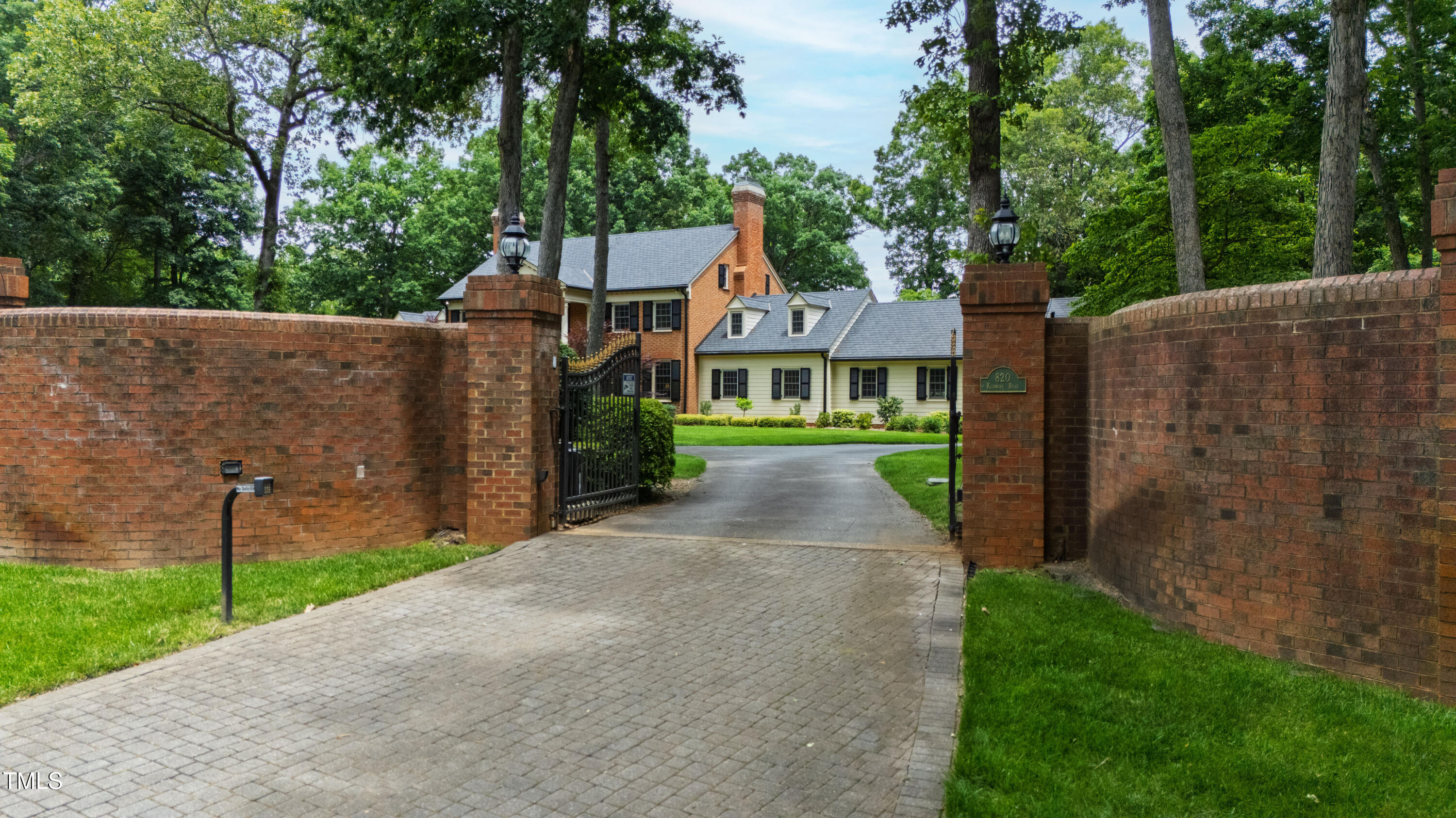 Brick home beyond an open iron gate, framed by brick walls and mature trees.