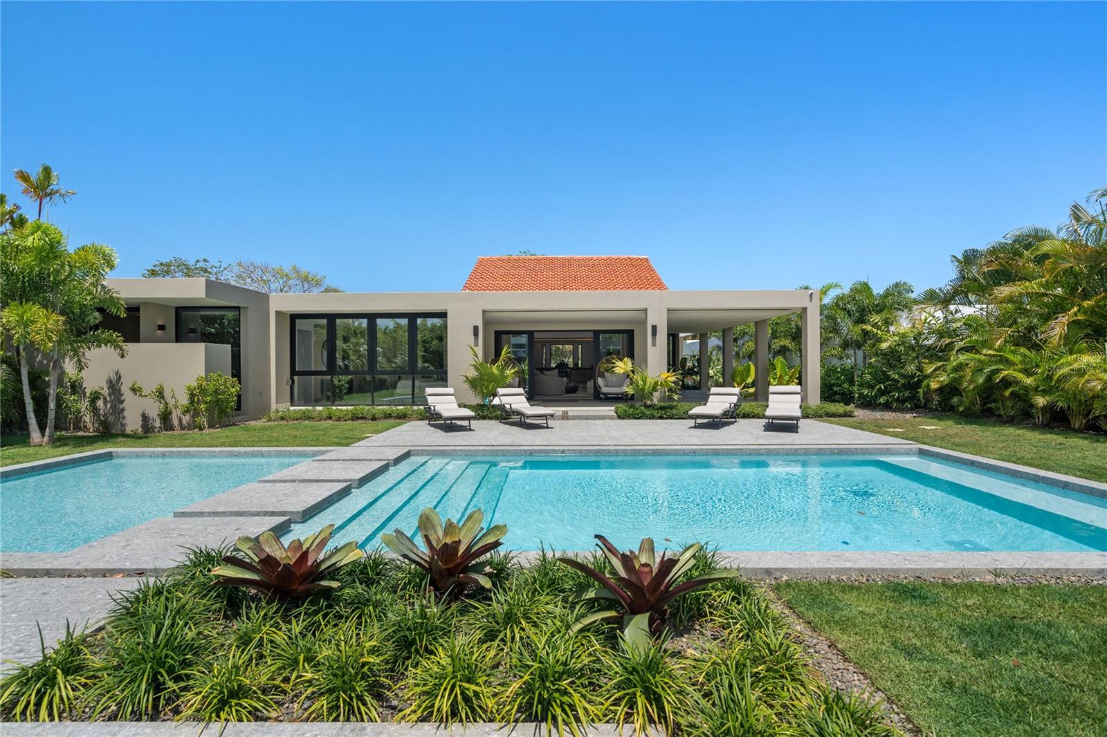 Modern home behind a turquoise pool, with broad entry steps, tropical landscaping, and patio loungers.
