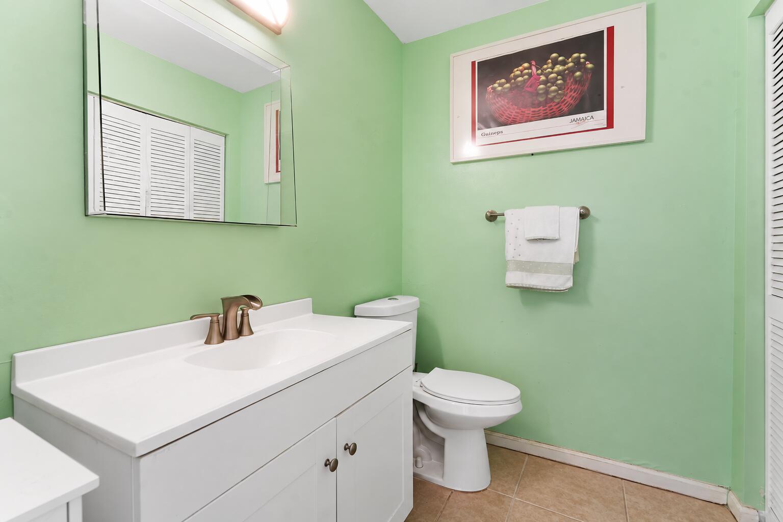 Bathroom with a white vanity and toilet against mint-green walls, plus a large mirror and framed artwork.