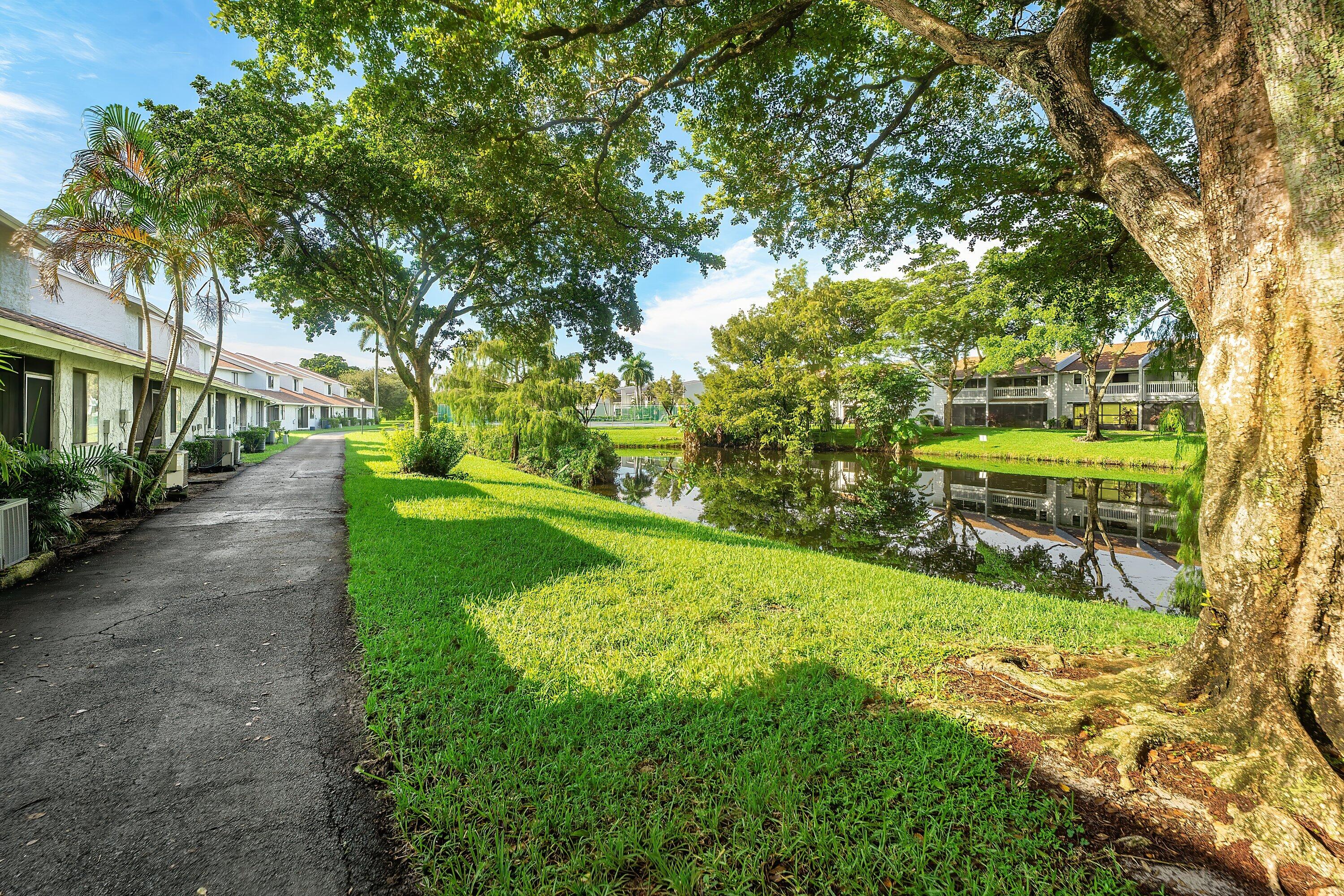 Residential community grounds with a pond, paved walkway, and broad shade trees.
