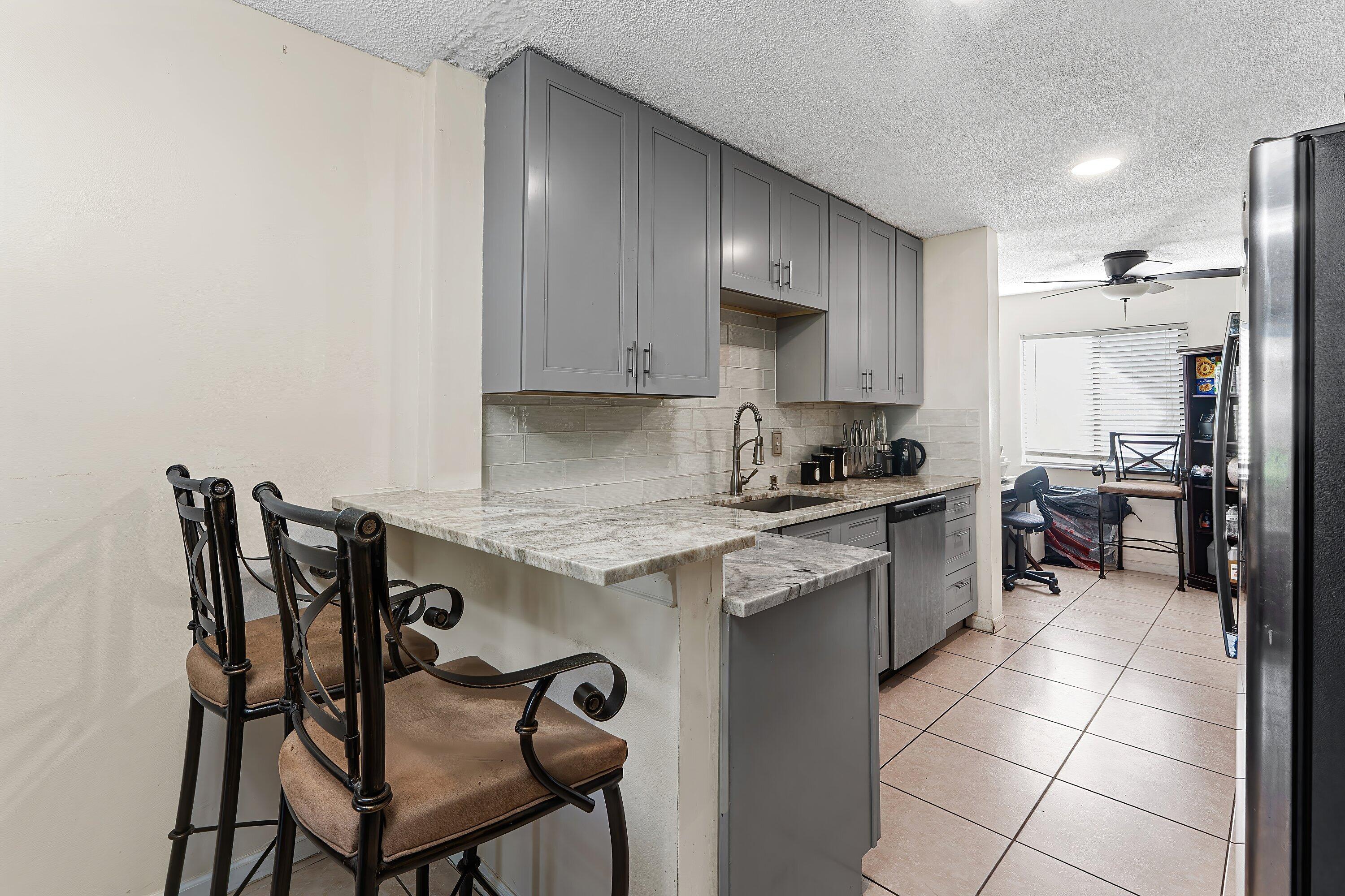 Kitchen with gray cabinets, stone breakfast bar and two chairs, opening onto a small dining area.