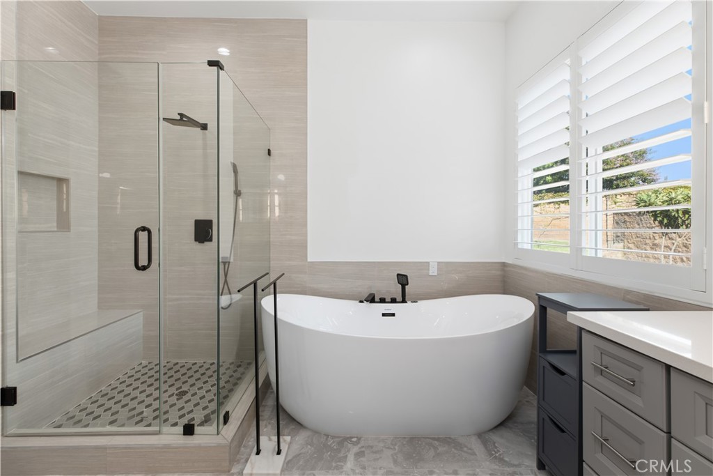 Modern bathroom with a glass shower, freestanding tub, and gray vanity beside shuttered windows.