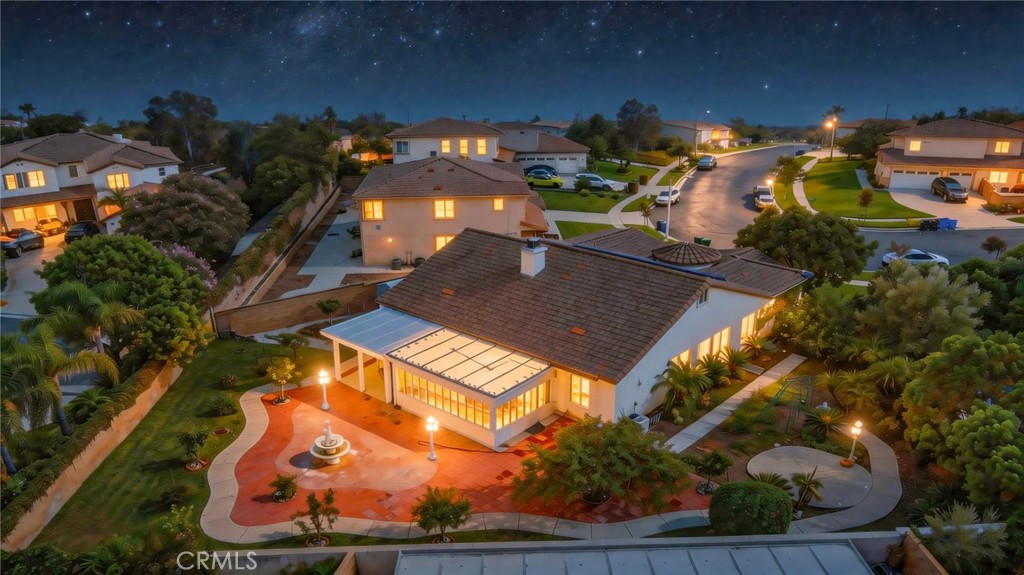 Aerial nighttime view of a tiled-roof home, landscaped patio, fountain, and surrounding neighborhood.