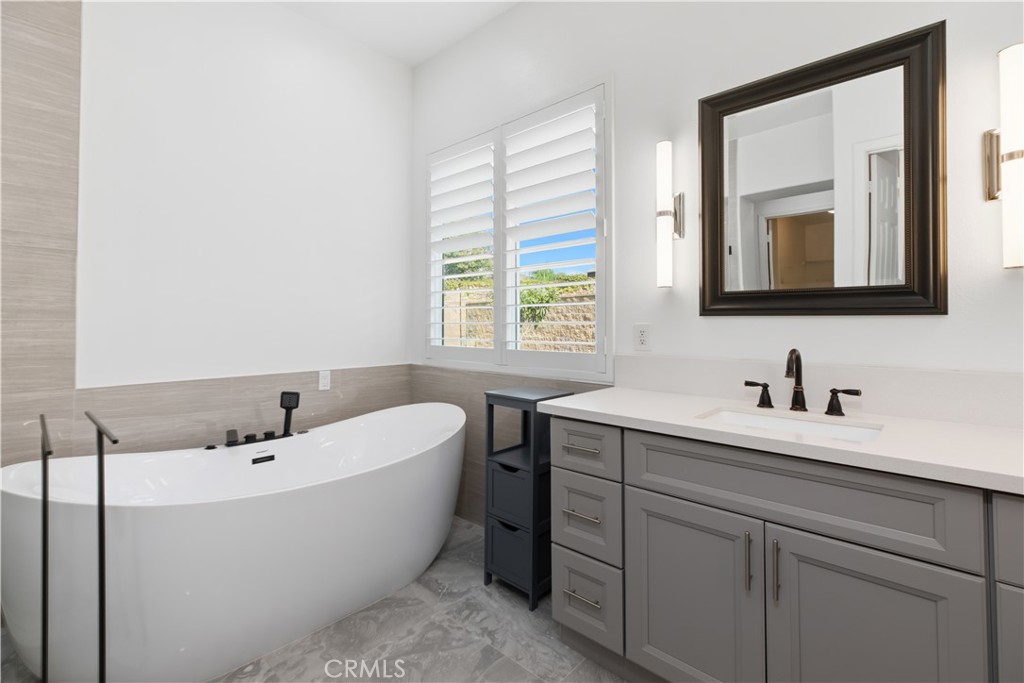 Bathroom with a freestanding soaking tub, gray vanity, and plantation-shuttered window.