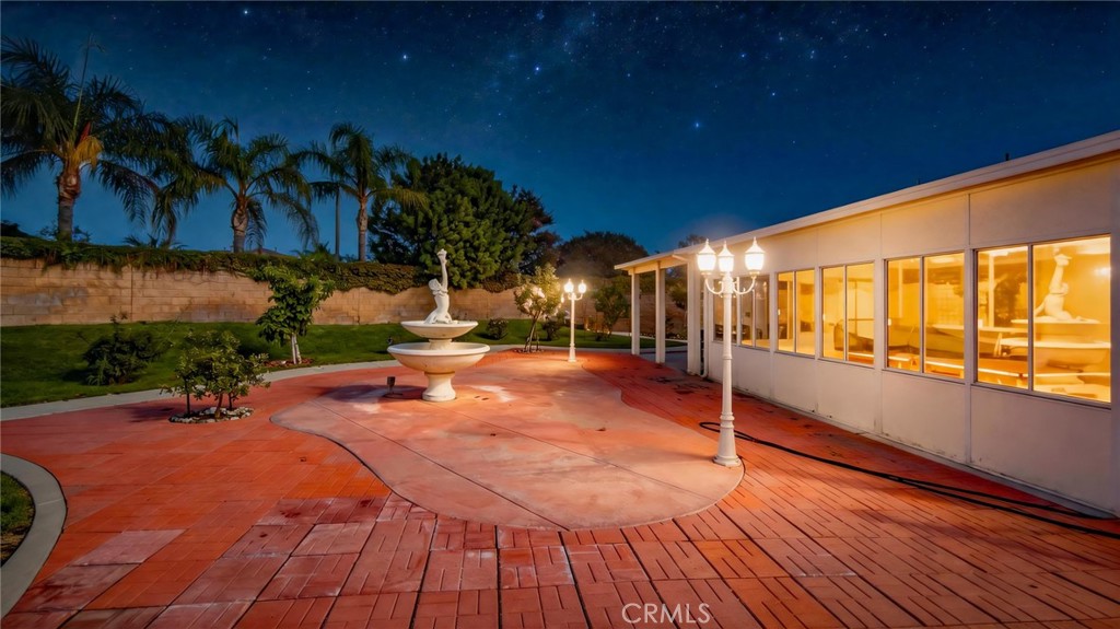 Backyard courtyard with a tiered fountain, red pavers, palm trees, and warmly lit house windows at night.