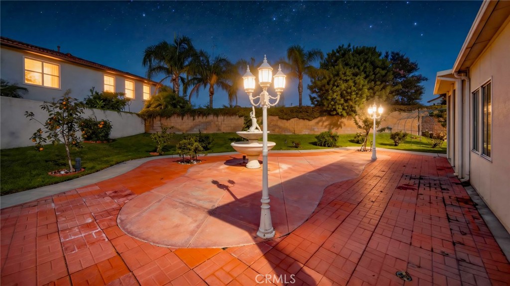 Backyard patio with red brick paving, lawn, palm trees, and glowing decorative lamps at night.