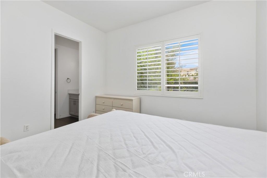 Bedroom with a bed, light dresser, and white plantation shutters beside an open bathroom doorway.