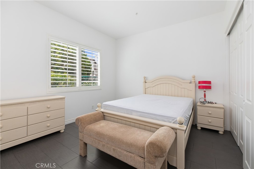 Bedroom with a cream bed, beige bench, dark tile floor, and shuttered window.