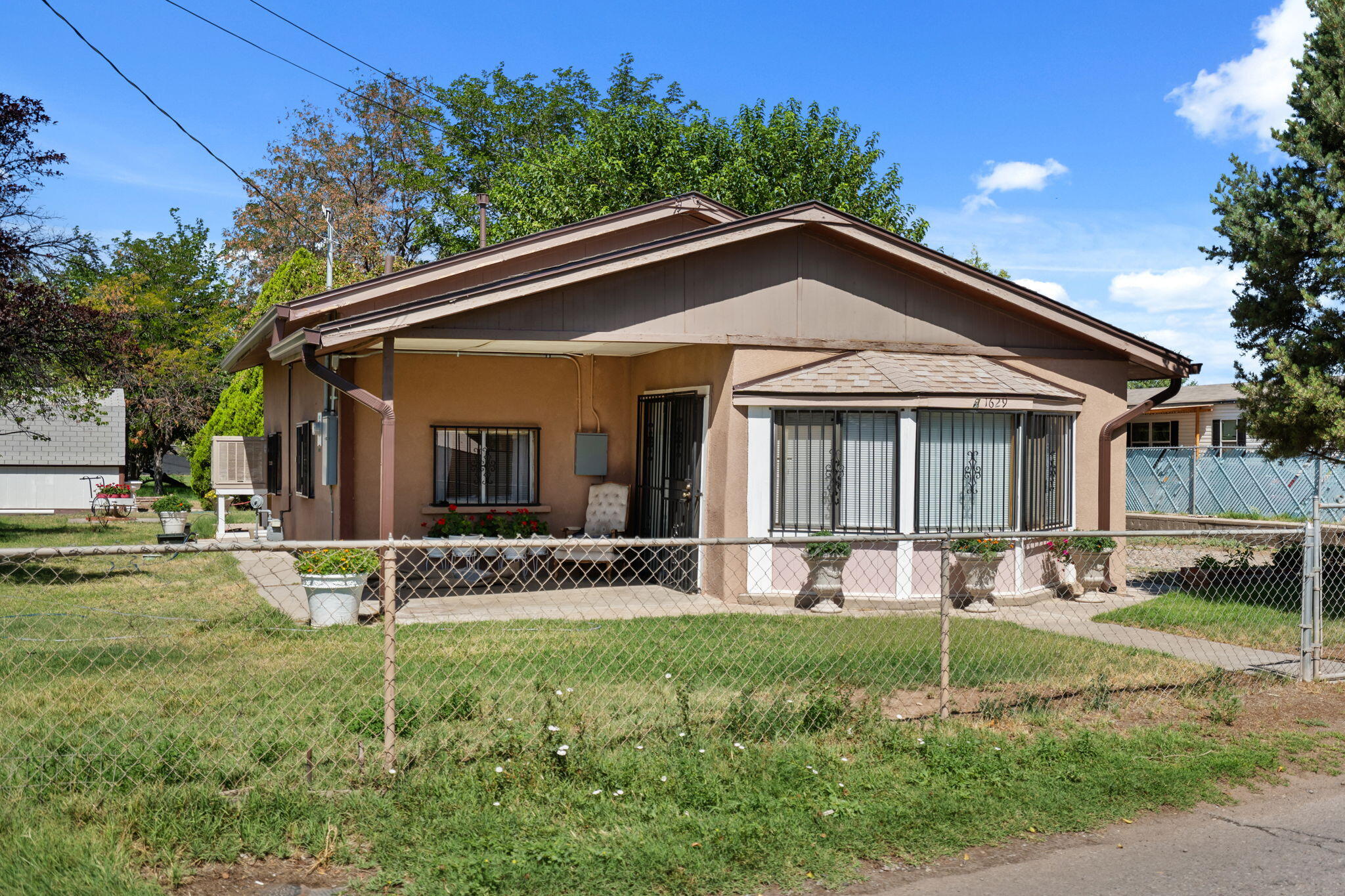 Single-story tan stucco house with a broad gabled roof, covered porch, and fenced front yard.