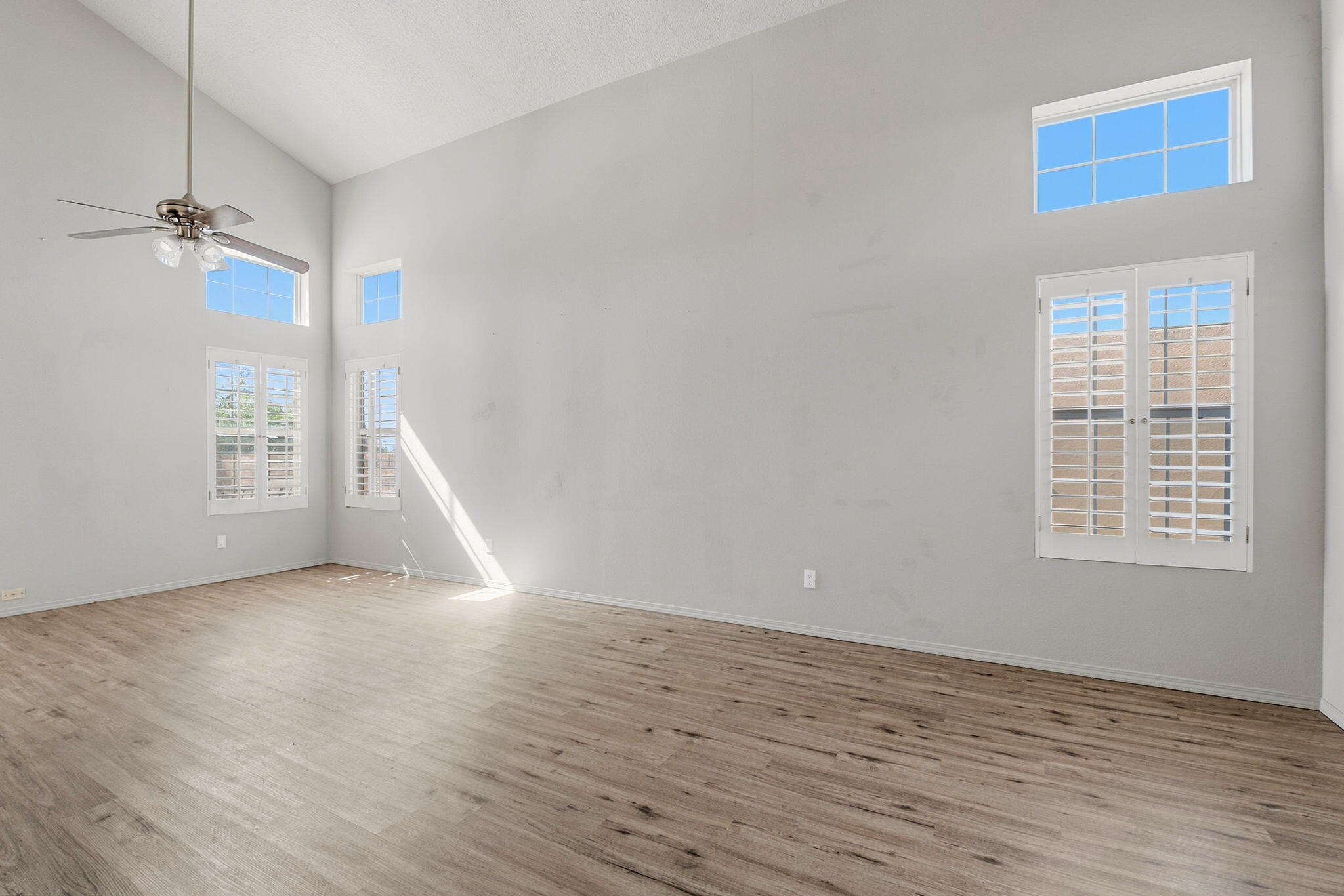 Empty living room with vaulted ceiling, wood-look flooring, ceiling fan, and shuttered windows.