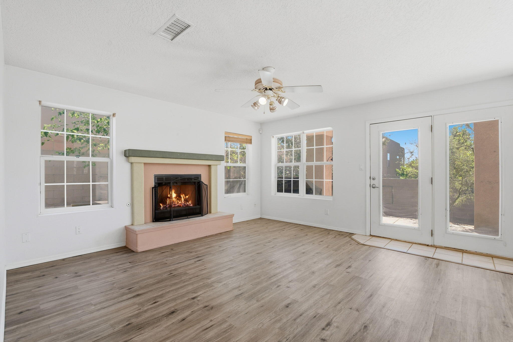 Empty living room with a lit fireplace, gray wood-look flooring, and windows beside glass doors.