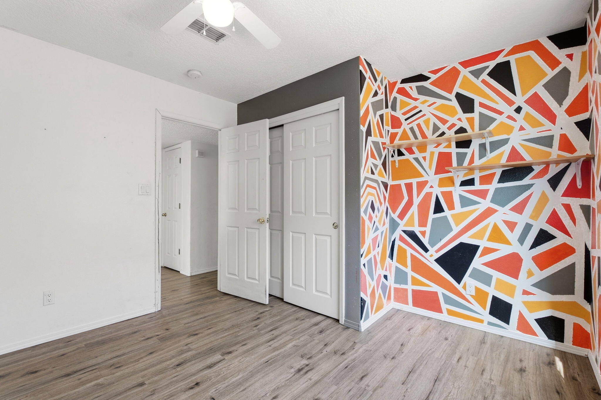 Empty bedroom with a colorful geometric accent wall, two wooden shelves, and open closet doors.