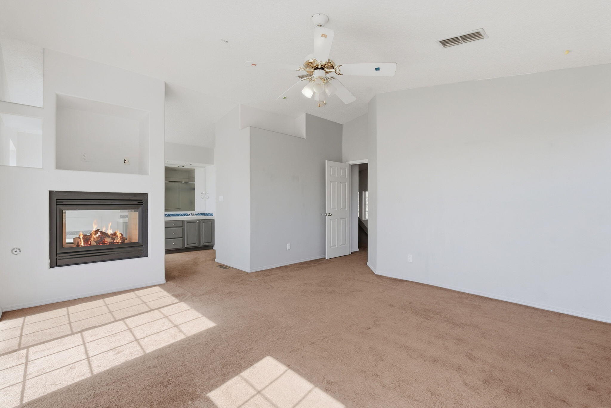 Empty living room with a lit fireplace, ceiling fan, and sunlit beige carpet.