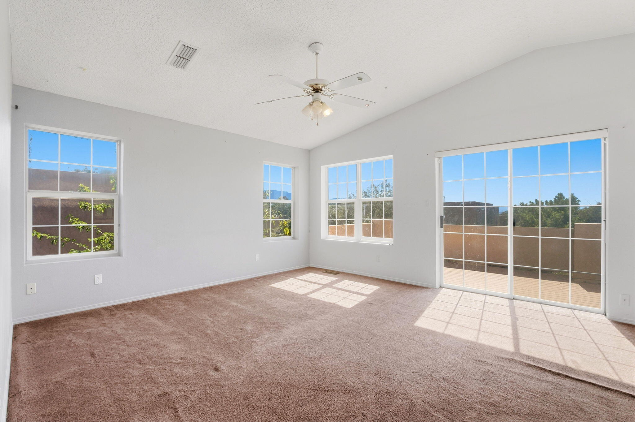 Empty carpeted room with a ceiling fan and sunlit windows beside a sliding glass door.