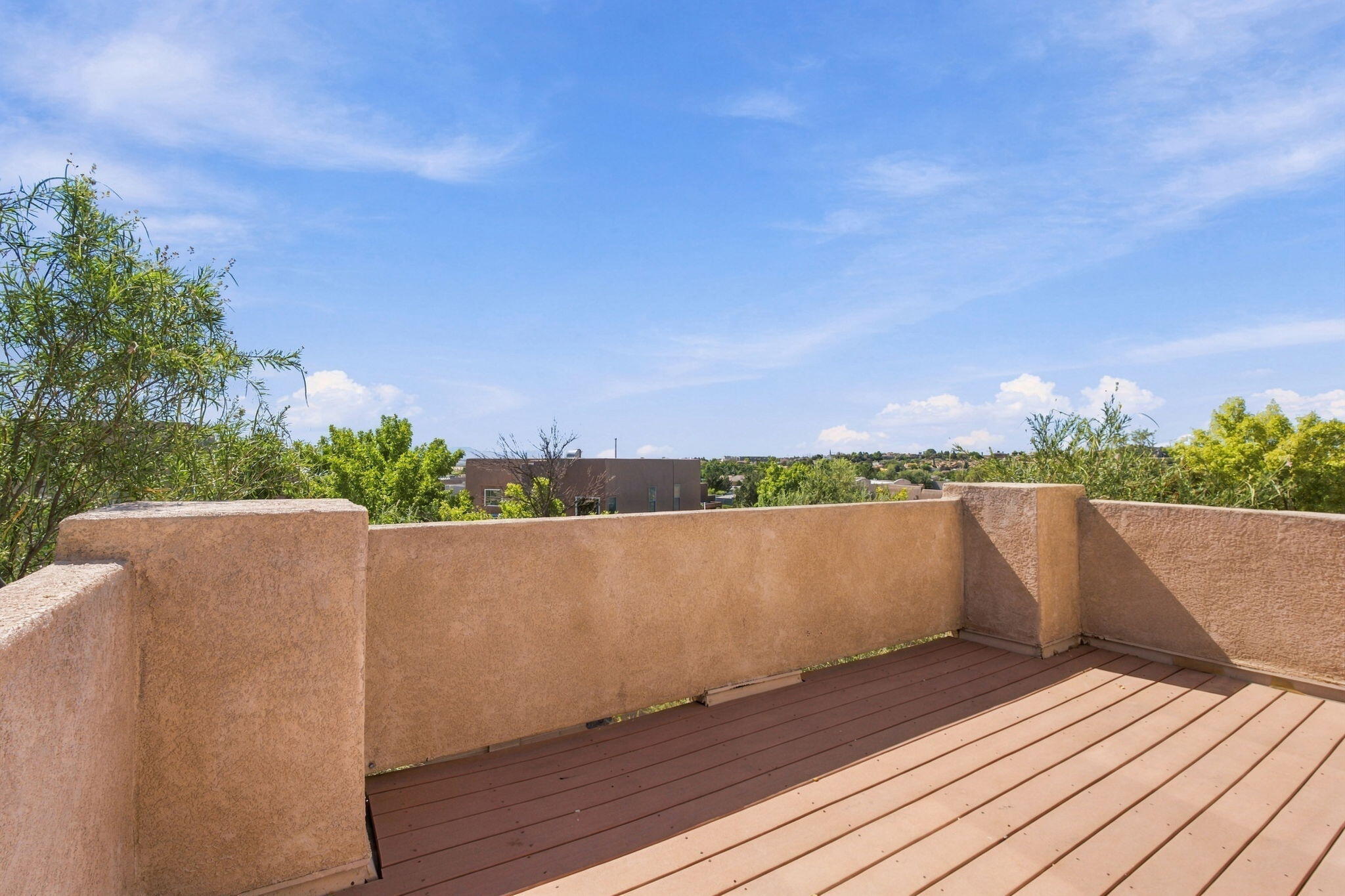 Outdoor deck with brown plank flooring, tan stucco walls, and trees and homes beyond.