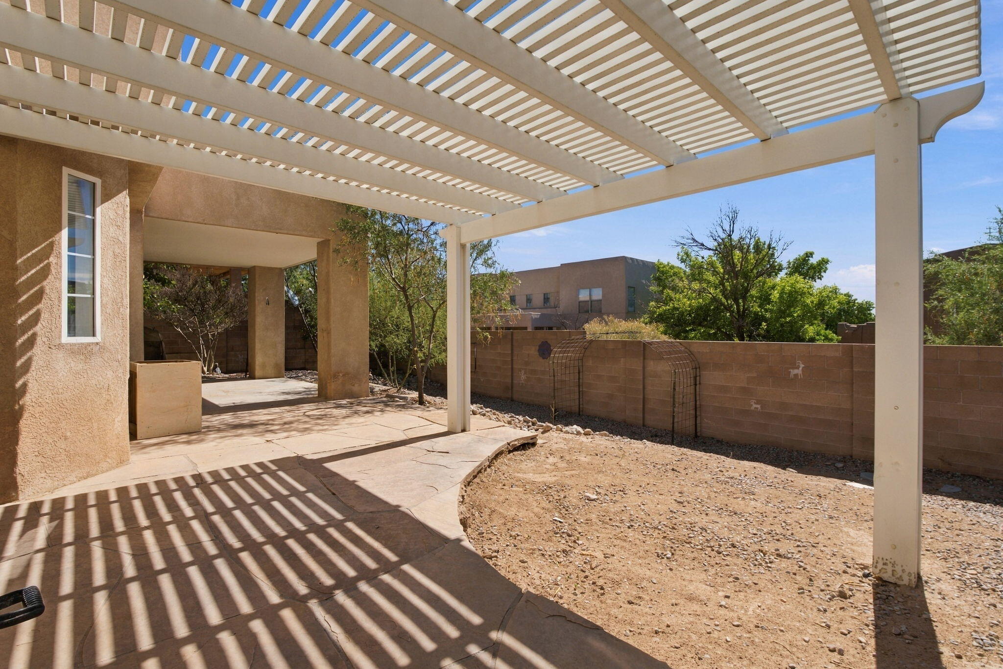 Backyard patio beneath a large white pergola, beside a dry gravel yard and block wall.